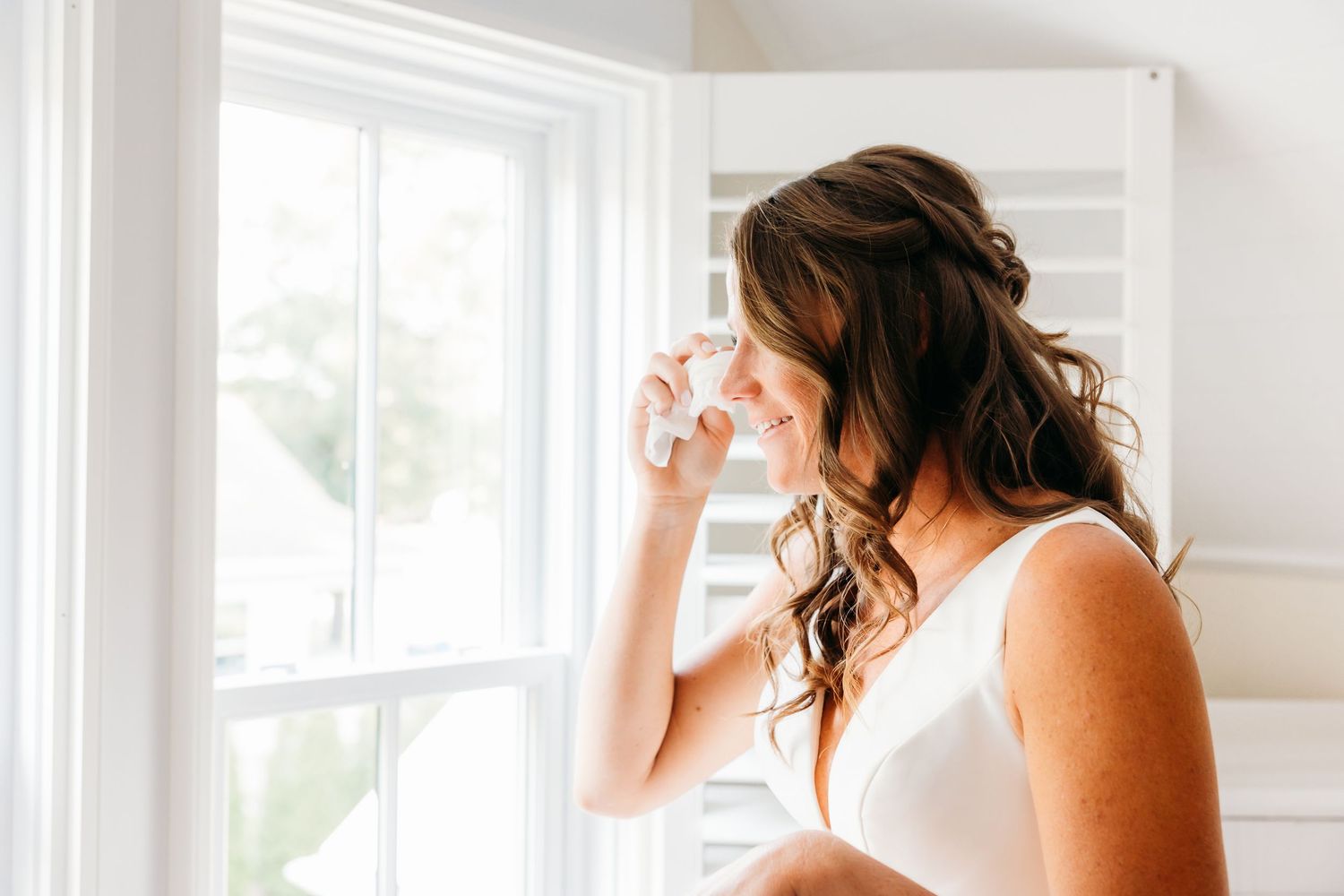 A person in a white dress stands near bright windows in an elegant room with natural lighting.