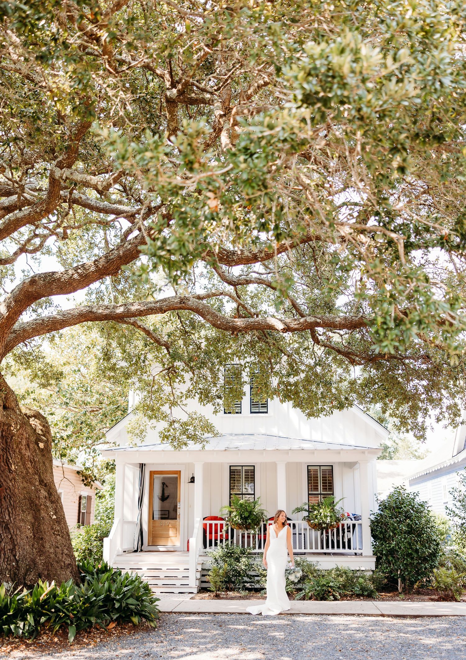 A white coastal cottage with wraparound porch is nestled under sprawling oak trees in a serene setting.