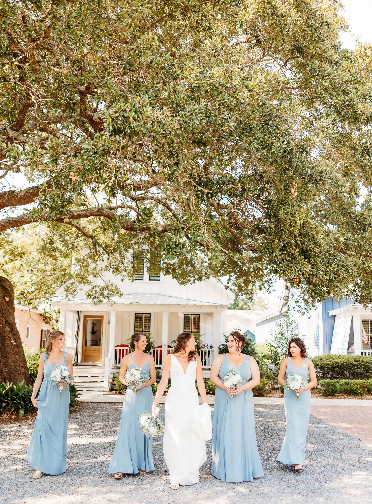A wedding party in light blue bridesmaid dresses poses together on a sunny path under oak trees at an outdoor venue.