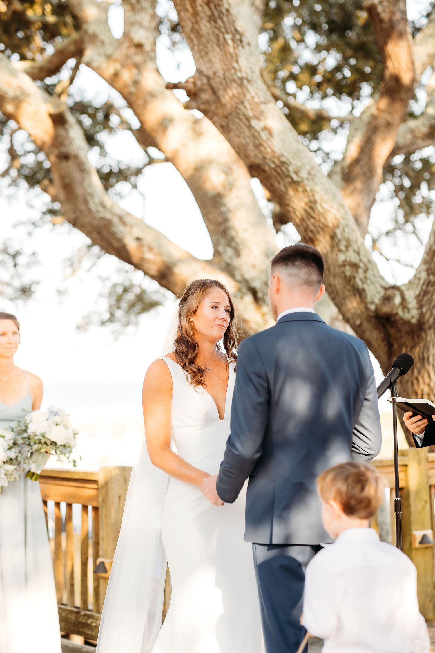 A couple shares a romantic moment during their outdoor wedding ceremony under large oak trees.