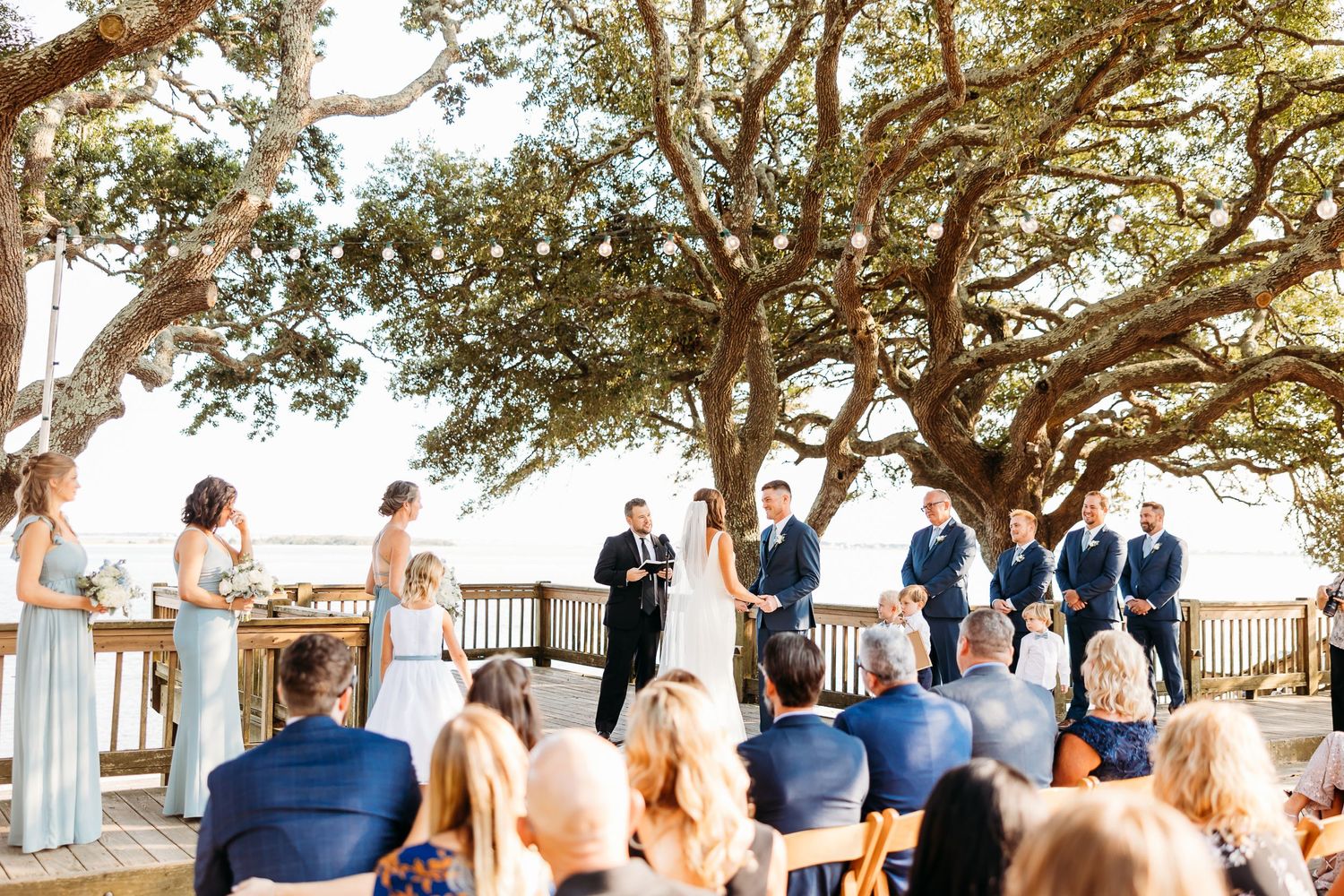Outdoor wedding ceremony under a majestic oak tree with guests seated on a wooden deck overlooking scenic views.