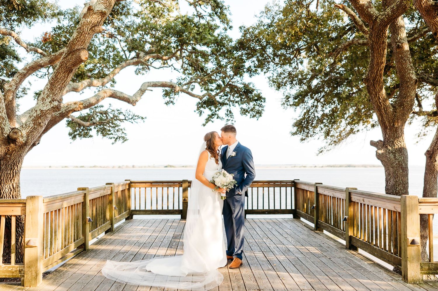 A romantic couple embraces on a wooden oceanfront boardwalk surrounded by trees during sunset.