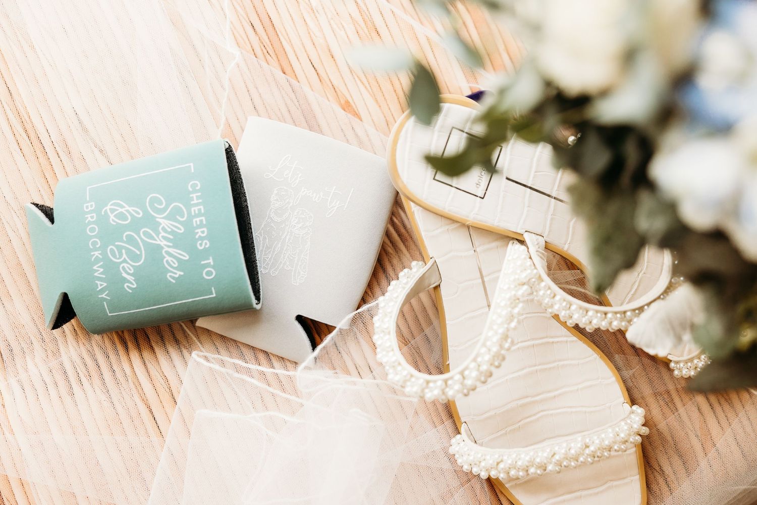 Wedding details featuring mint green koozies and white bridal sandals arranged on a peach-colored surface with eucalyptus.