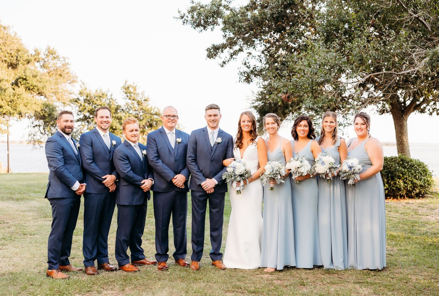 Wedding party in navy suits and light blue dresses poses together on a sunny outdoor lawn.