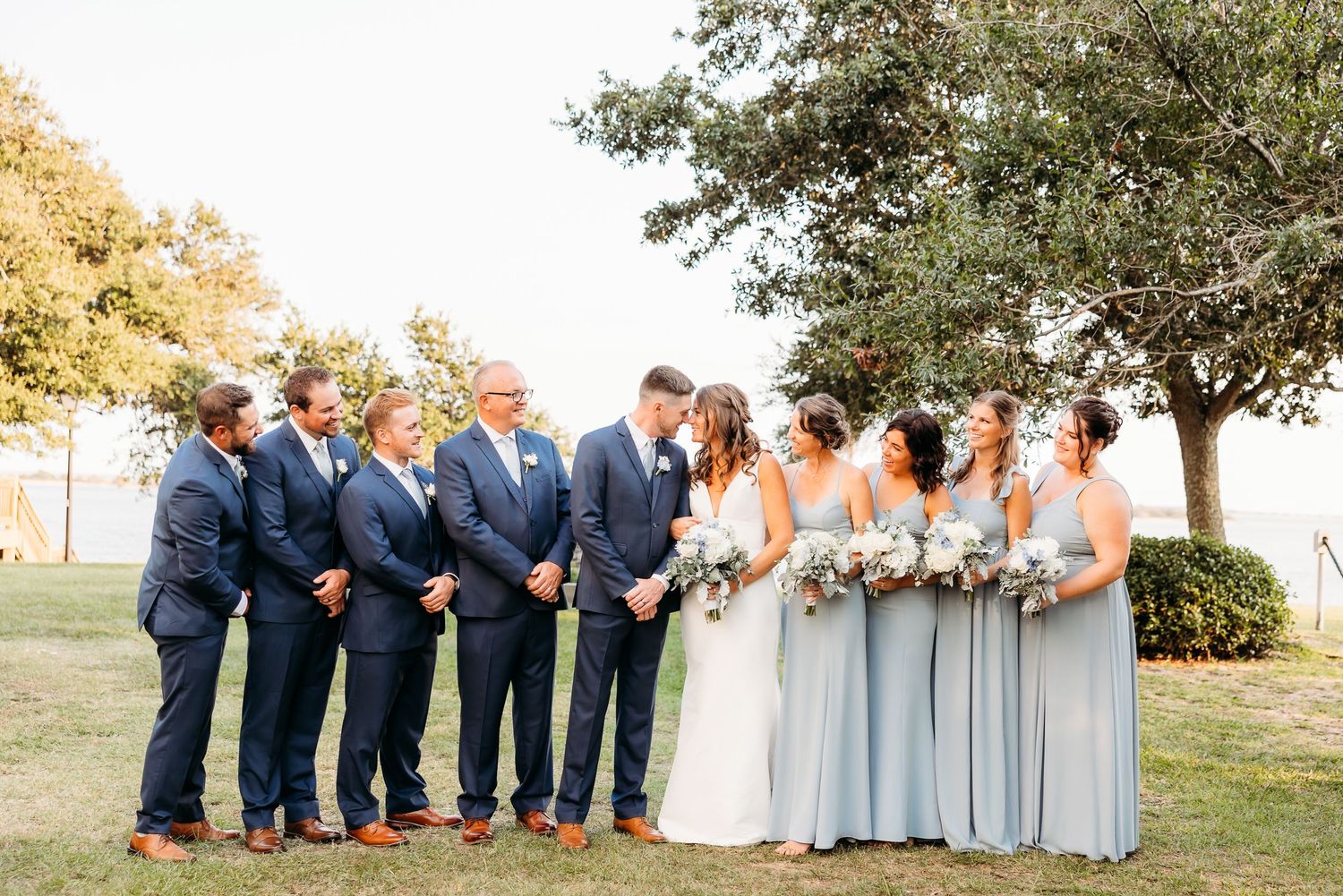 Wedding party in navy blue and light blue attire poses together outdoors under a tree during golden hour.