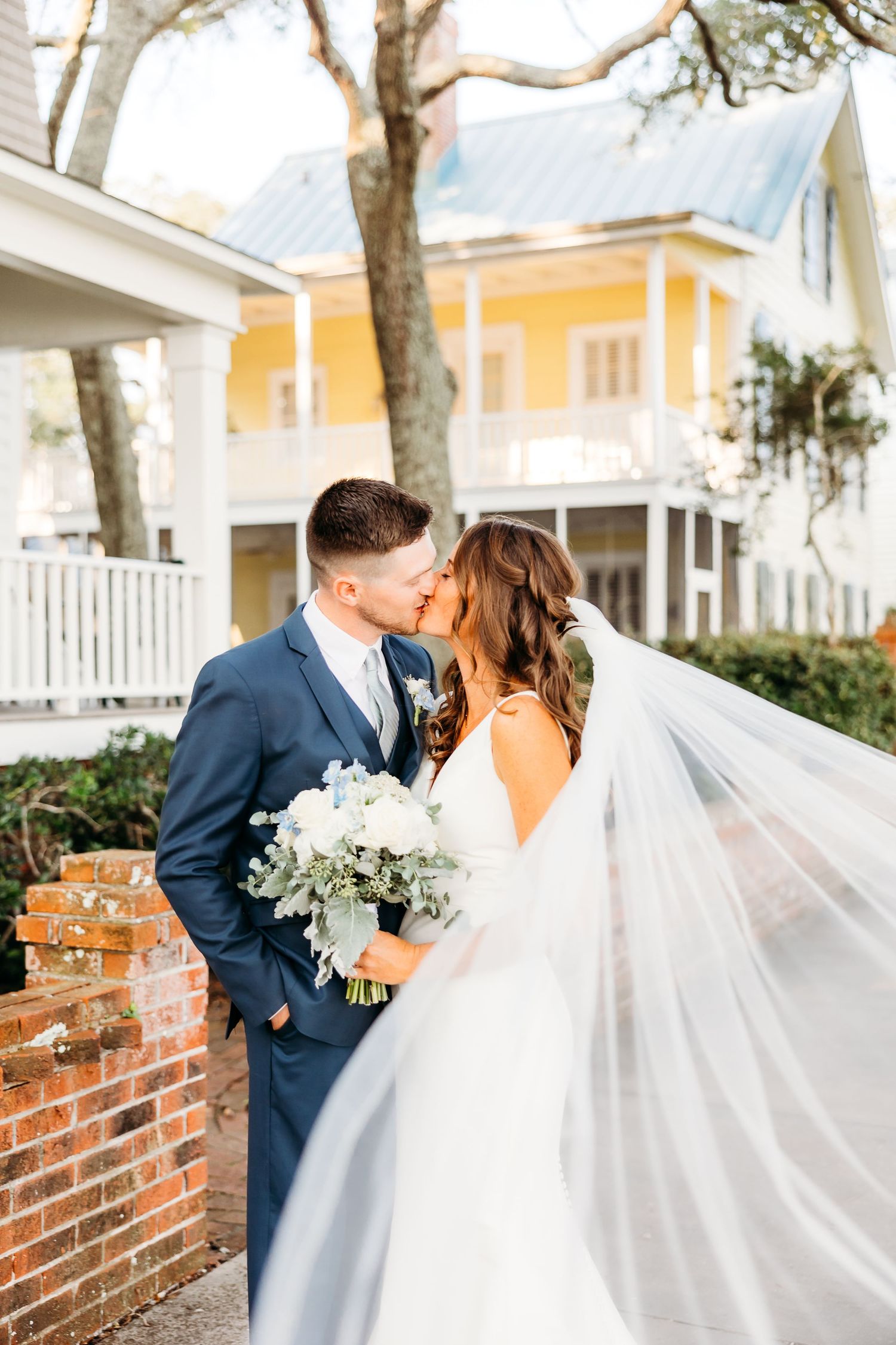 A romantic wedding couple shares a tender moment on a brick walkway with a flowing white veil in the breeze.