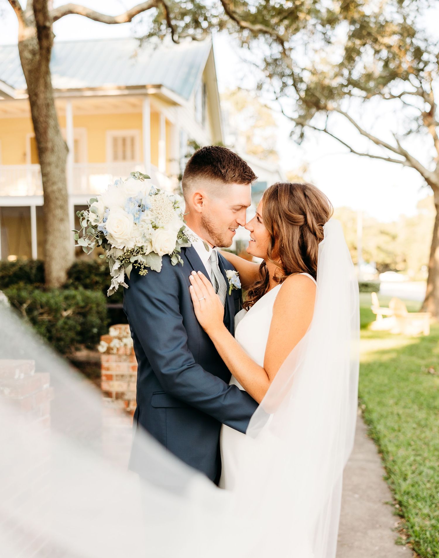 A romantic wedding couple shares tender kisses in a garden setting with trees and a yellow building in the background.