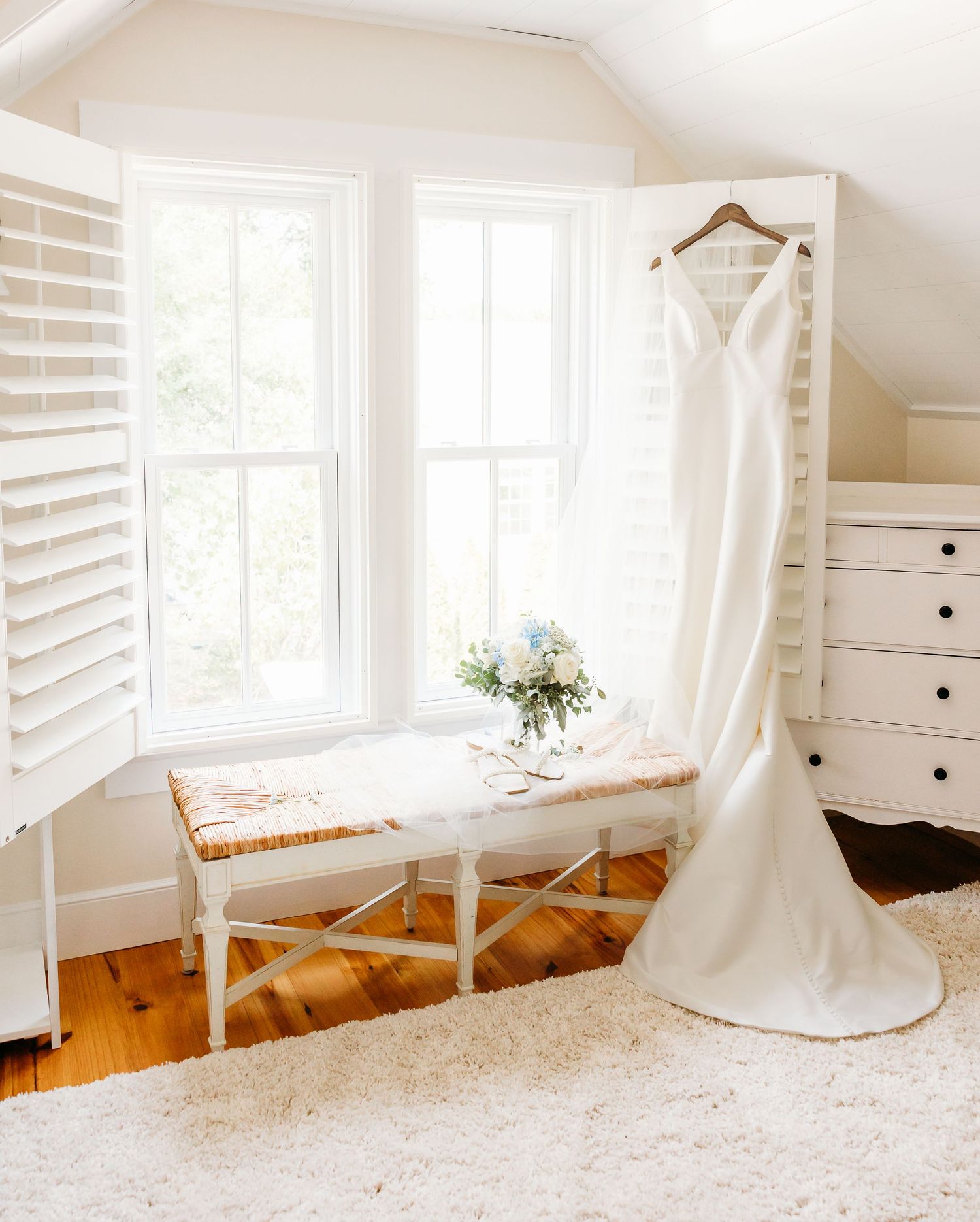 A bright white wedding dress hangs in a sunlit room with white shutters and warm hardwood floors.