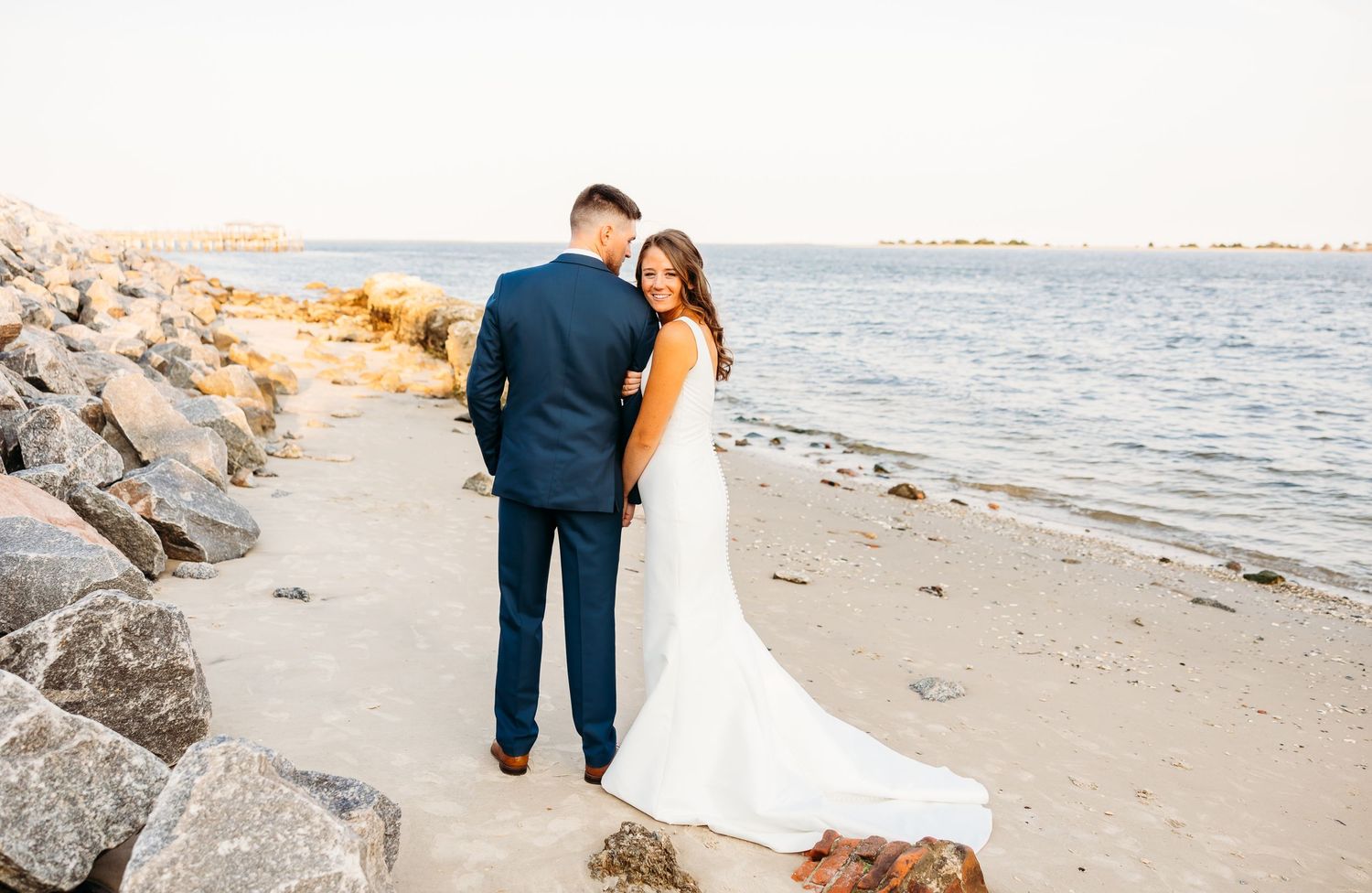 A couple in wedding attire walks along a rocky beach at sunset, creating a romantic coastal scene.