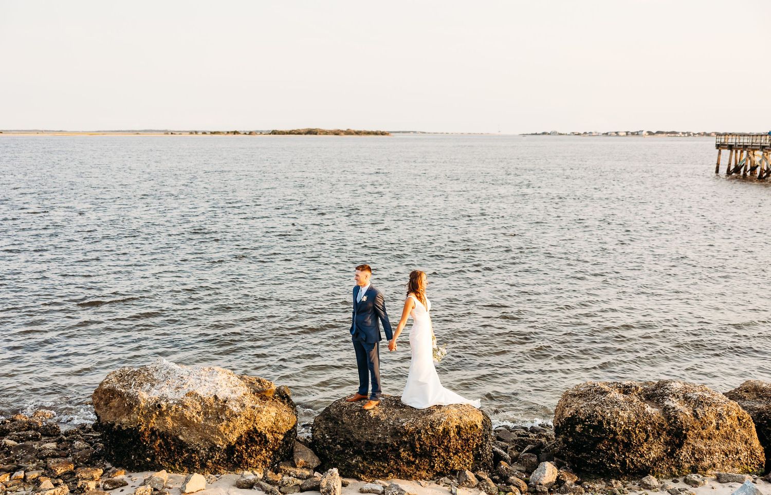 A couple in wedding attire stands on rocky coastal cliffs overlooking a serene ocean at sunset.