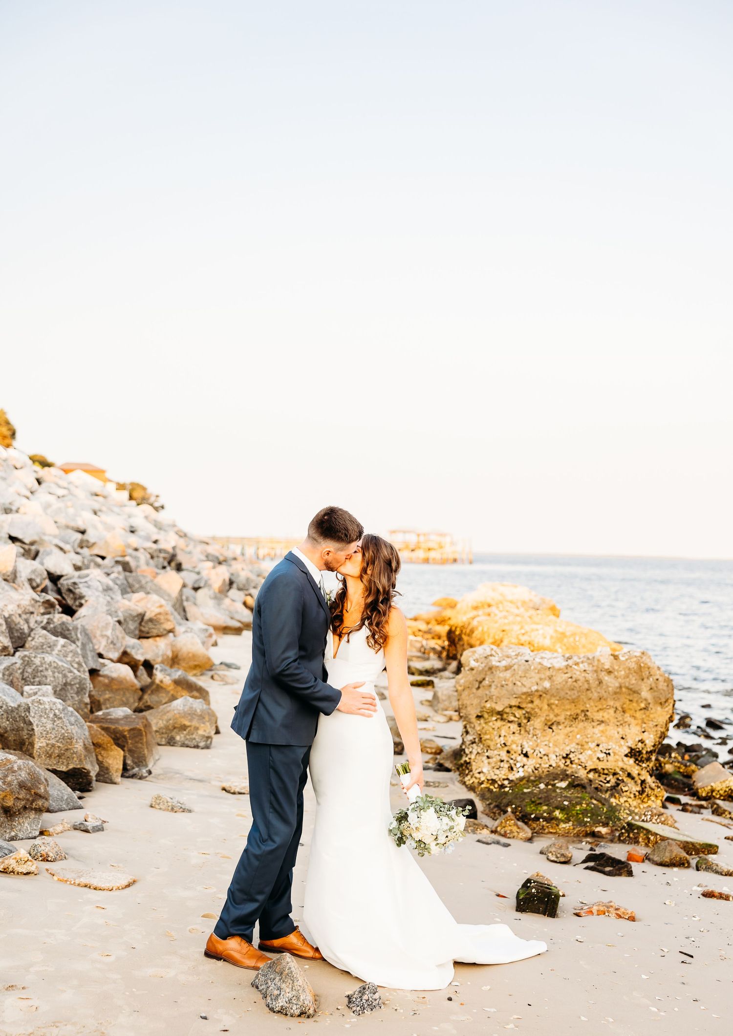 A couple in wedding attire shares an intimate moment on a rocky beach at sunset.