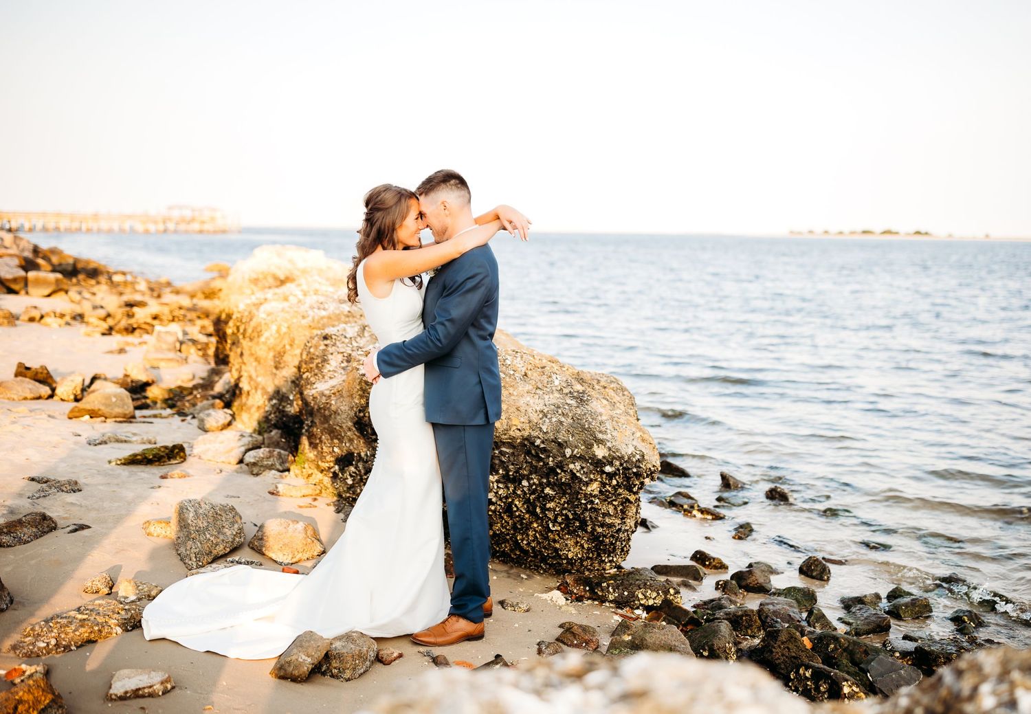 A couple shares an intimate moment on a rocky beach at sunset with waves crashing in the background.