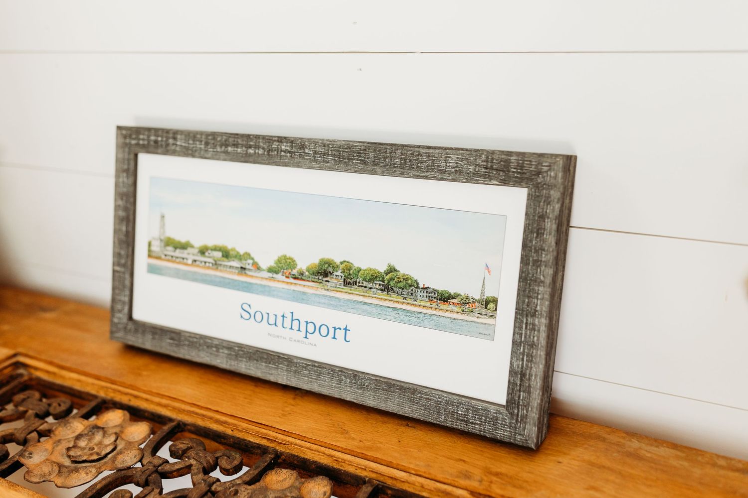 A series of framed artwork pieces displayed on a wooden shelf against a white wall.