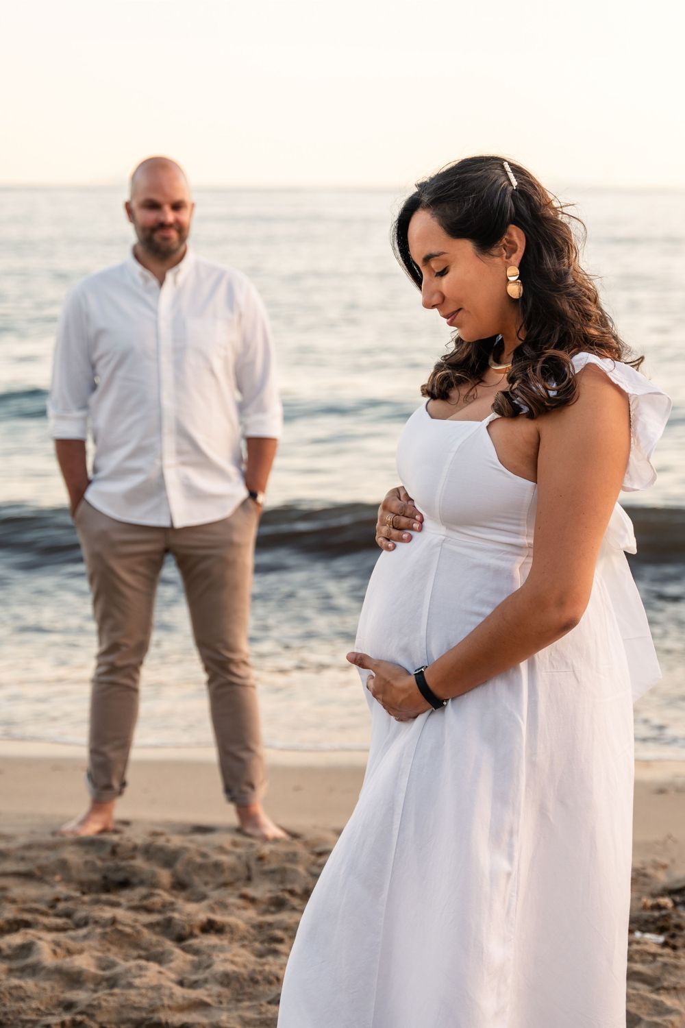 couple wearing white and neutral clothing on the beach for maternity photo shoot woman look at her pregnant belly