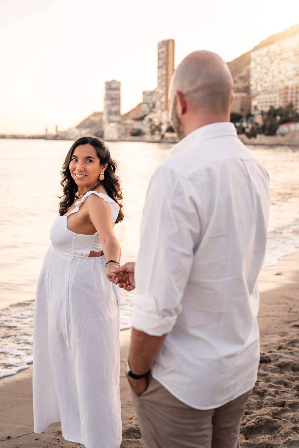 woman wearing white dress looking back at man and holding his hand walking towards the water on the beach at sunset
