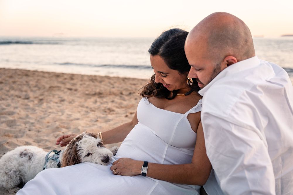 Couple cuddling and sitting on the sand on the beach and their dog resting his head on the lap of pregnant woman
