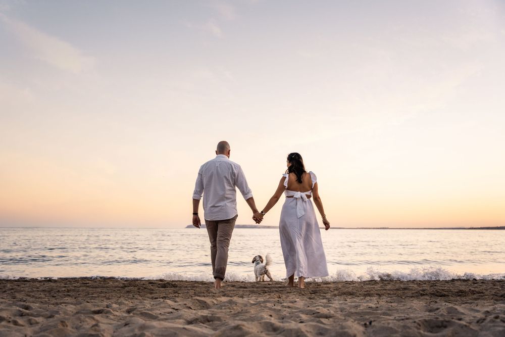 couple holding hands and walking towards the water at sunset with their dog in between them