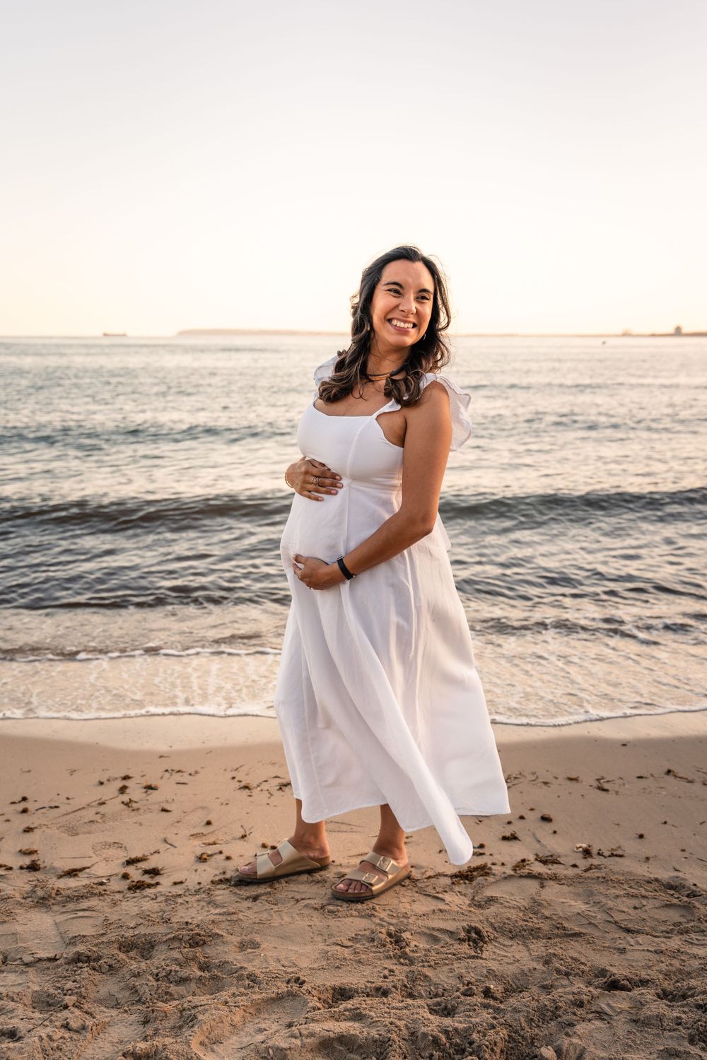 Pregnant woman wearing white dress on the beach at sunset holding her belly and smiling