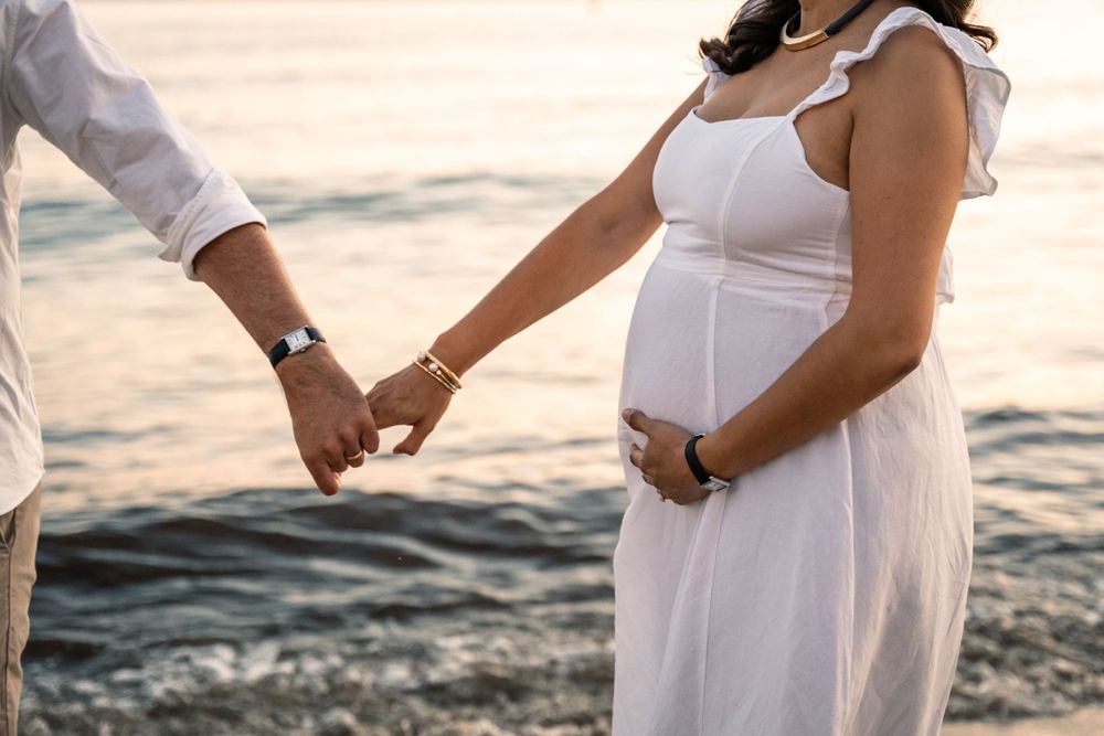 couple holding hands and woman holding her pregnant belly on the beach with soft light they are wearing white