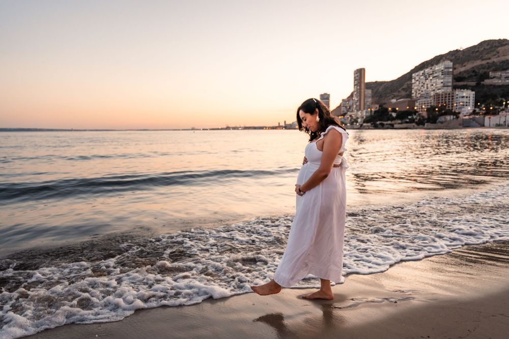pregnant woman wearing white dress on the beach at sunset walking towards the water and holding her pregnant belly