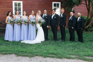 Wedding party in formal attire poses outdoors in front of red barn with bridesmaids in light purple dresses.