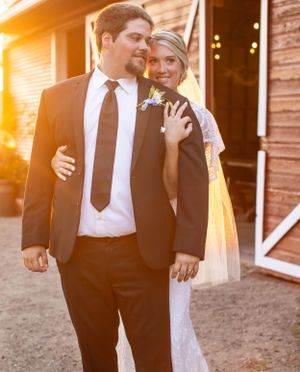 Sunset portrait of newlyweds in formal attire against rustic barn backdrop.