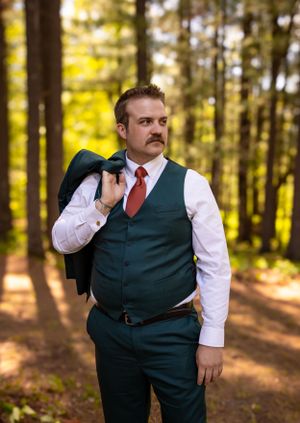 Groomsman in green vest and red tie posing in sunlit forest setting.