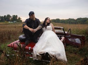 Wedding couple sitting on vintage car in rustic field setting.