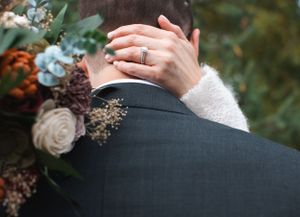 Close up of hands with wedding ring embracing against formal attire and floral bouquet.