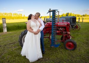Wedding couple posing near vintage tractors in a rustic farm field at sunset.