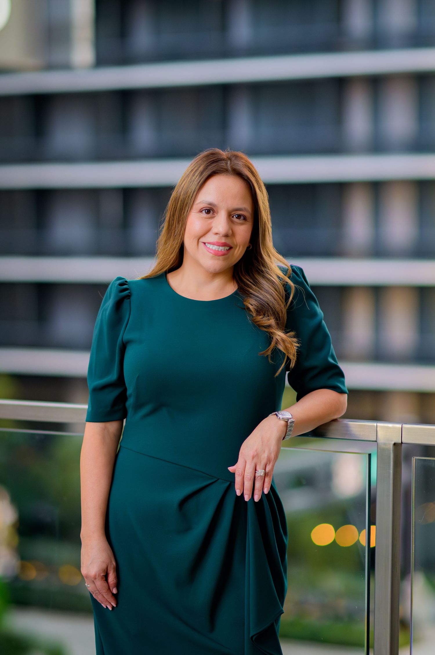 Professional in dark green dress stands elegantly by glass railing with city background.