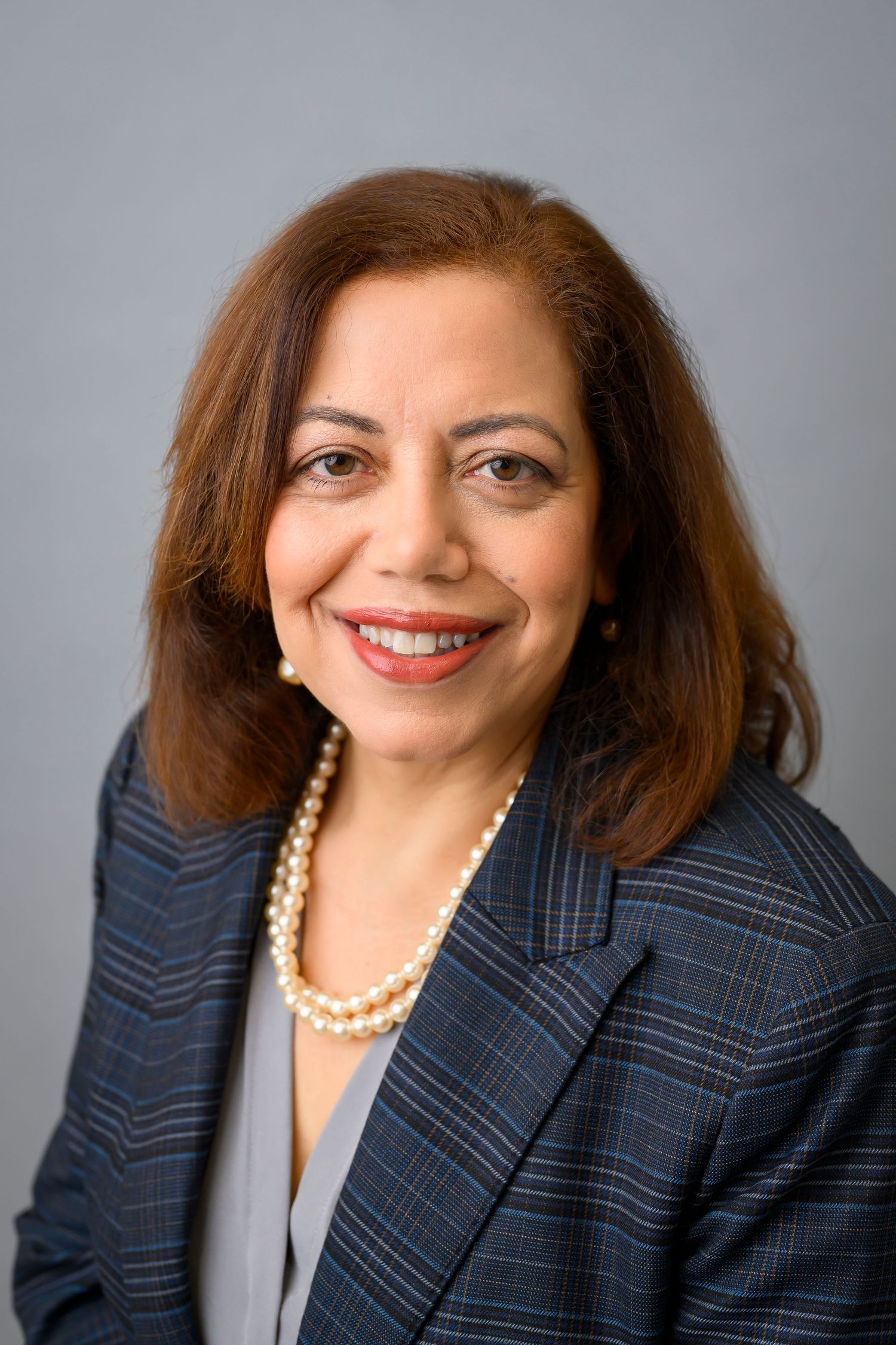 Professional headshot of woman in pinstriped suit and pearl necklace against gray background.