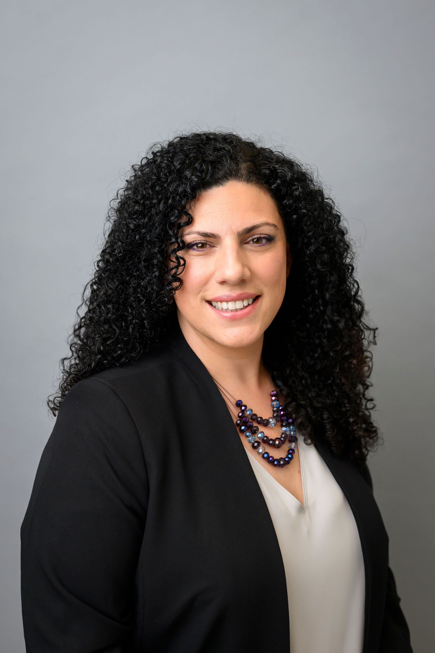 Professional headshot of a woman in black blazer against grey background.
