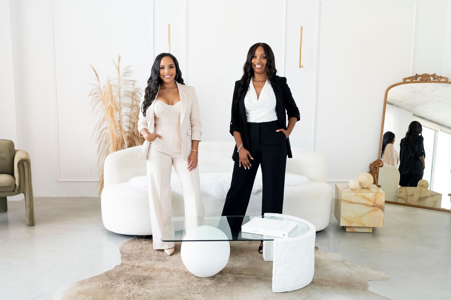 Two women in contrasting black and white outfits standing in modern white room with decorative elements.
