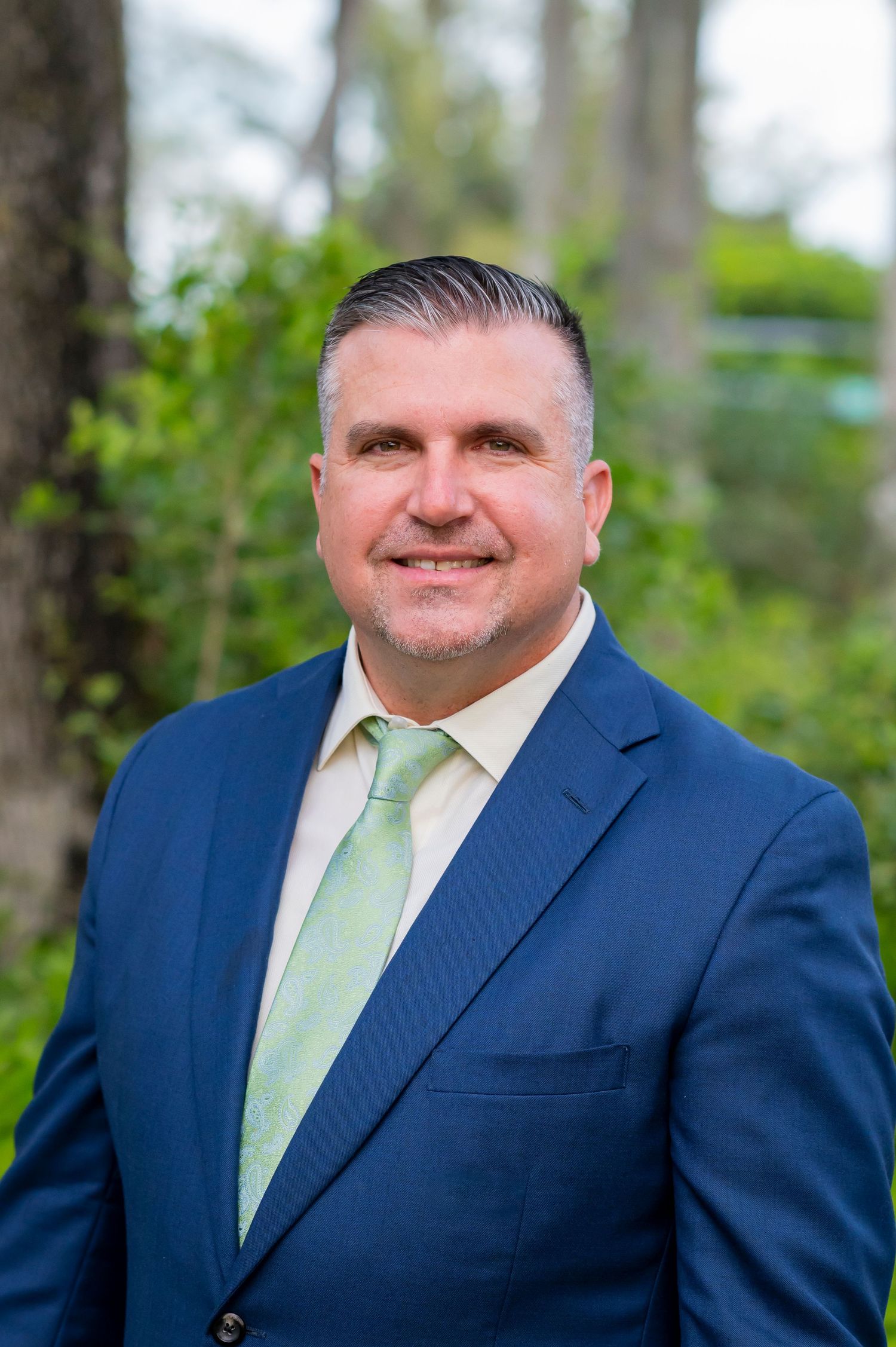 Professional headshot portrait with navy suit and mint green tie outdoors with natural background.