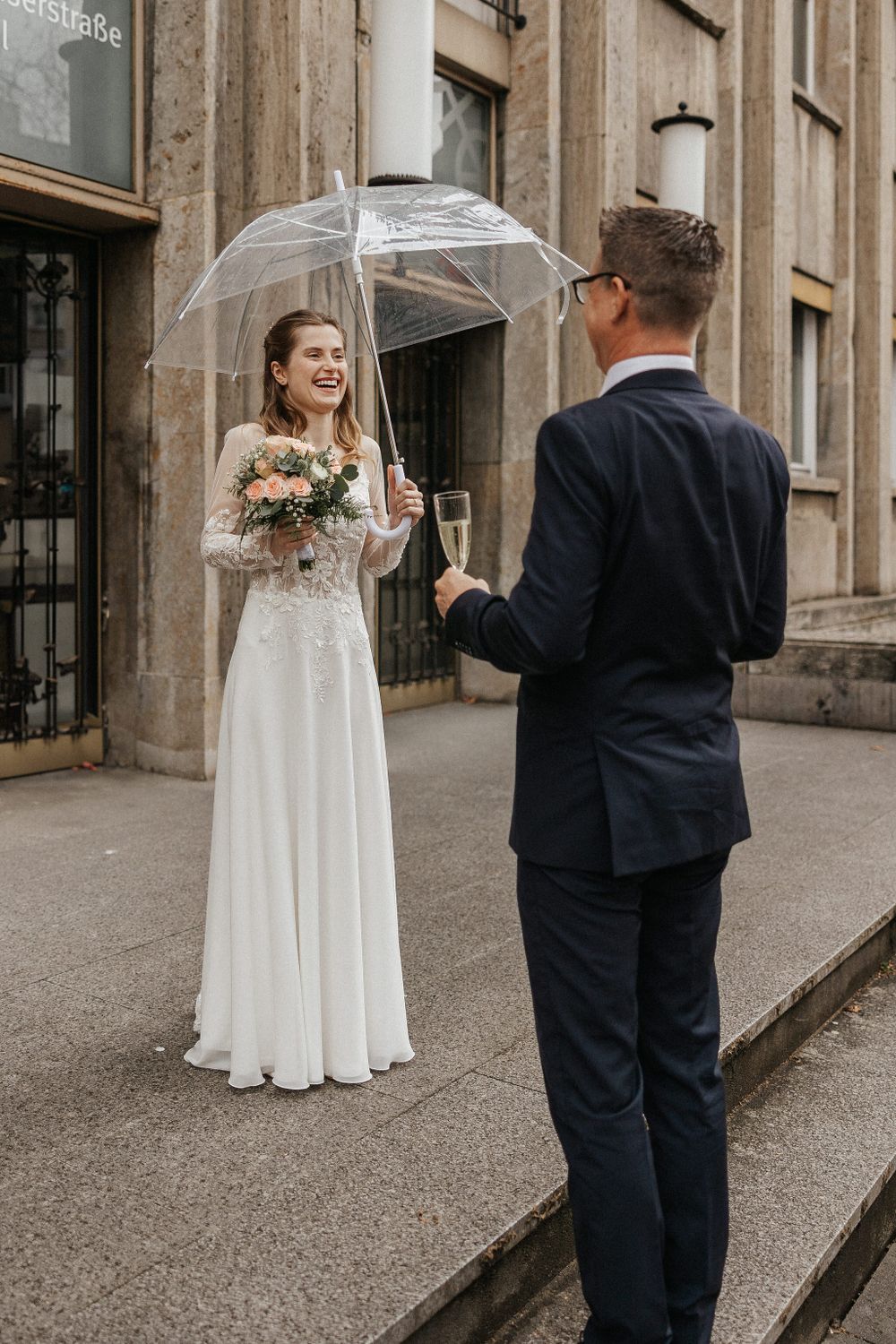 A couple shares champagne and laughter under a clear umbrella outside a historic building.
