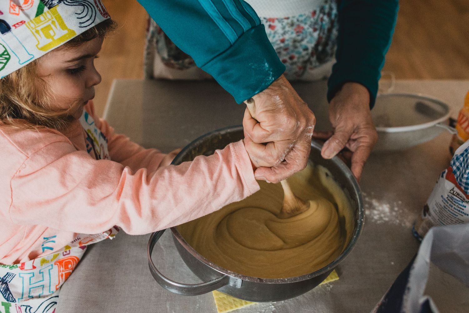 nieta con delantal de cocina y gorro revolviendo una mezcla, la no de su abuela ayudándola a revolver