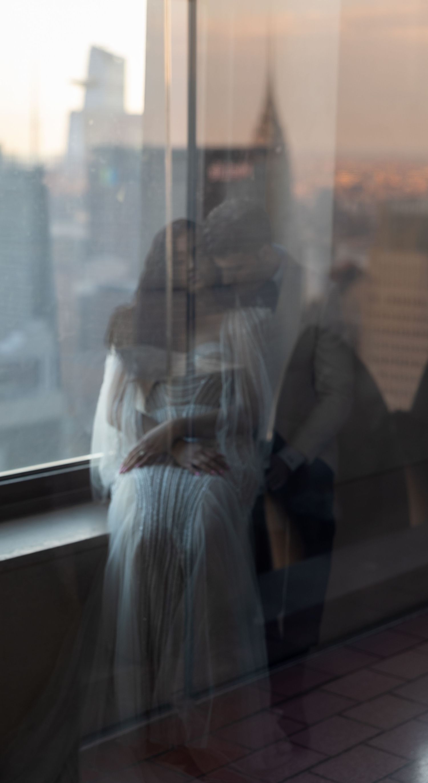 Reflective window view of couple in formal wear against city skyline at sunset.