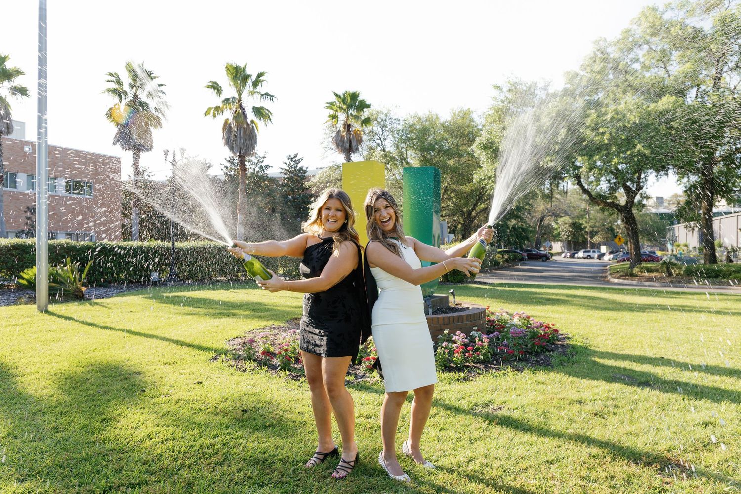 Two women celebrate outdoors by popping champagne bottles on a sunny green lawn with palm trees and colorful flowers.