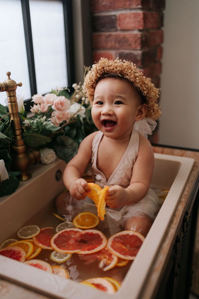 A joyful baby wearing a flower crown plays with citrus fruits in a vintage bathtub.