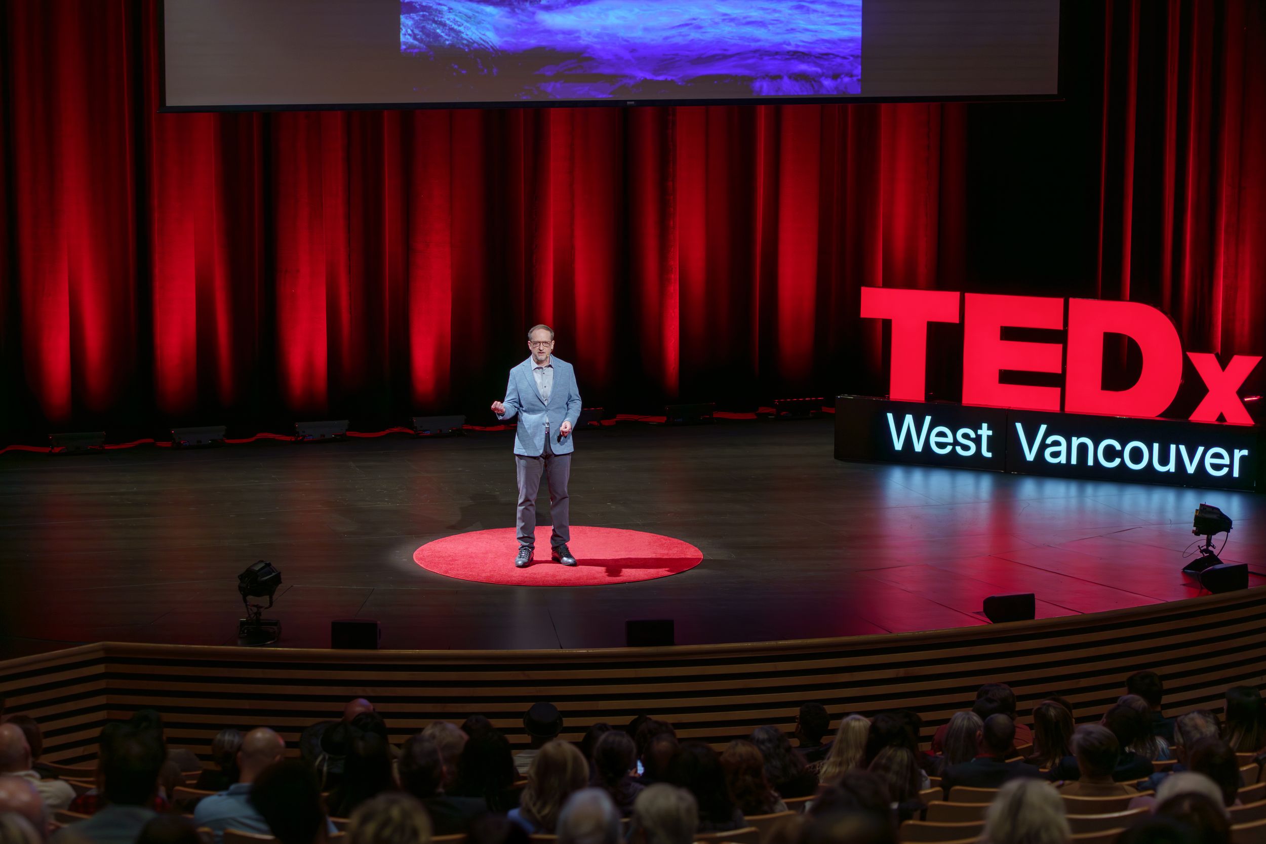 A speaker stands on a red circular stage at a TEDx West Vancouver event with dramatic red lighting and theater seating.
