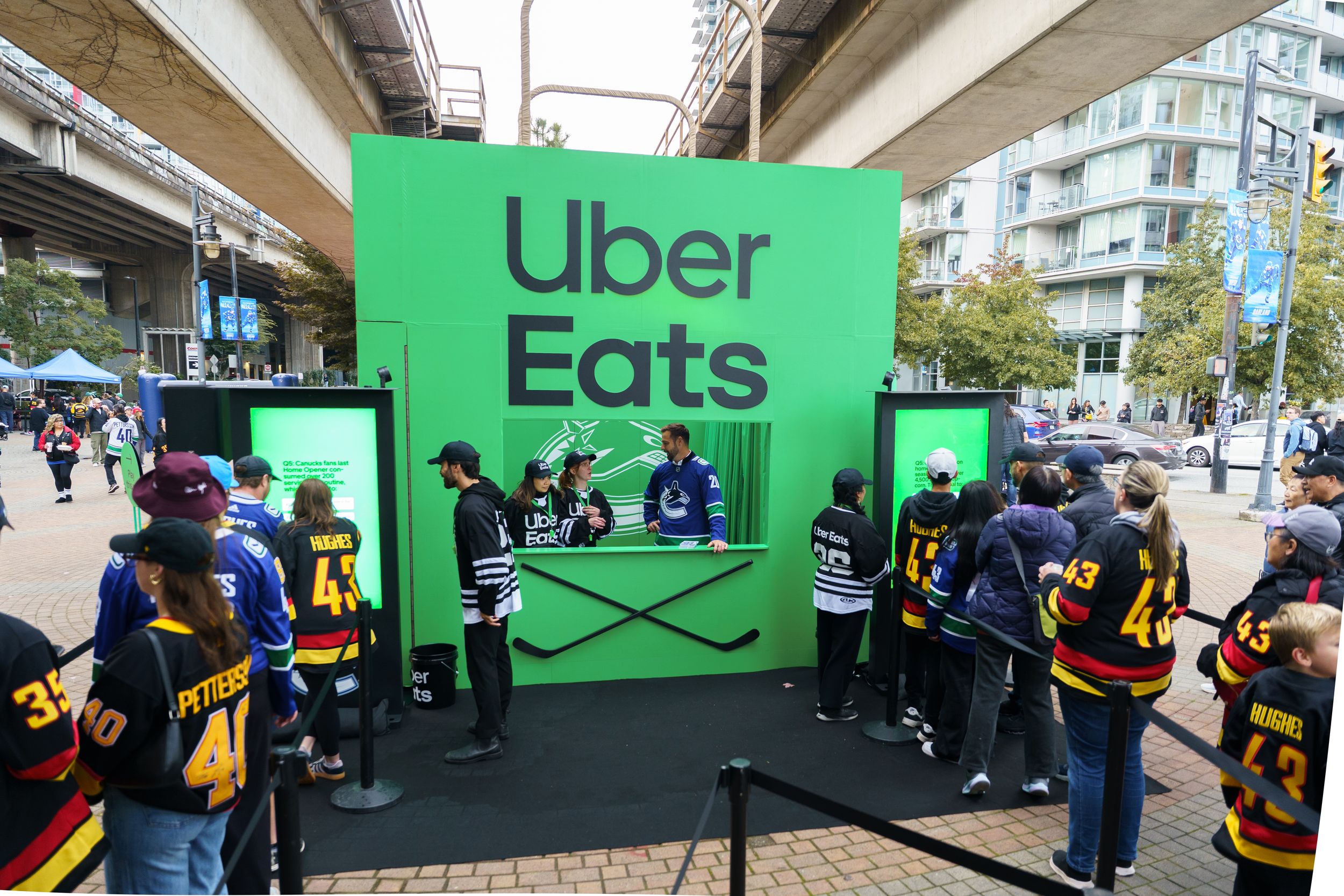 Food delivery workers in yellow bags wait in line at green Uber Eats pickup stations under an urban overpass.