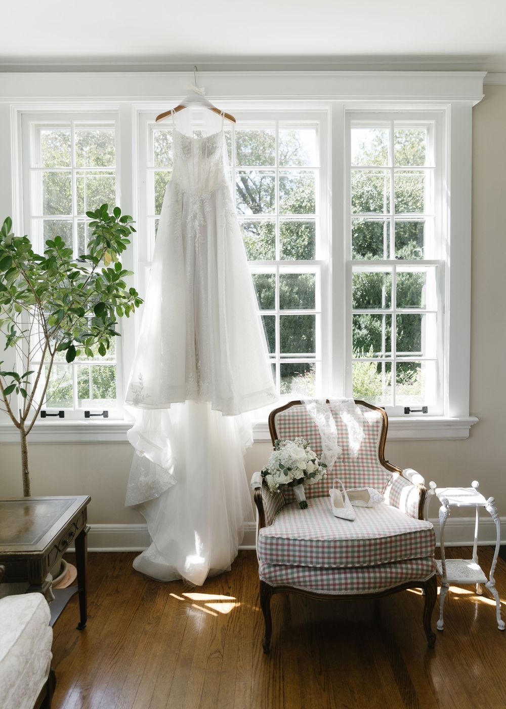 Bright sunlit room with white curtains, gingham chaise lounge, and potted plants by large windows.