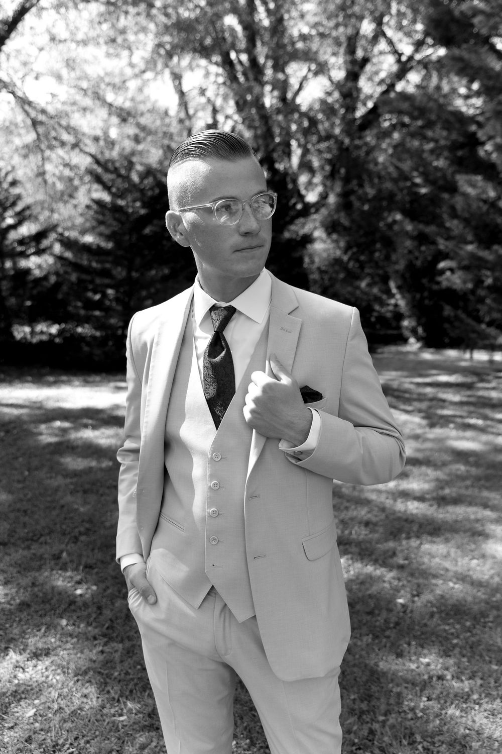 Close-up shots of a groom in a light colored three-piece suit with glasses in an outdoor setting.