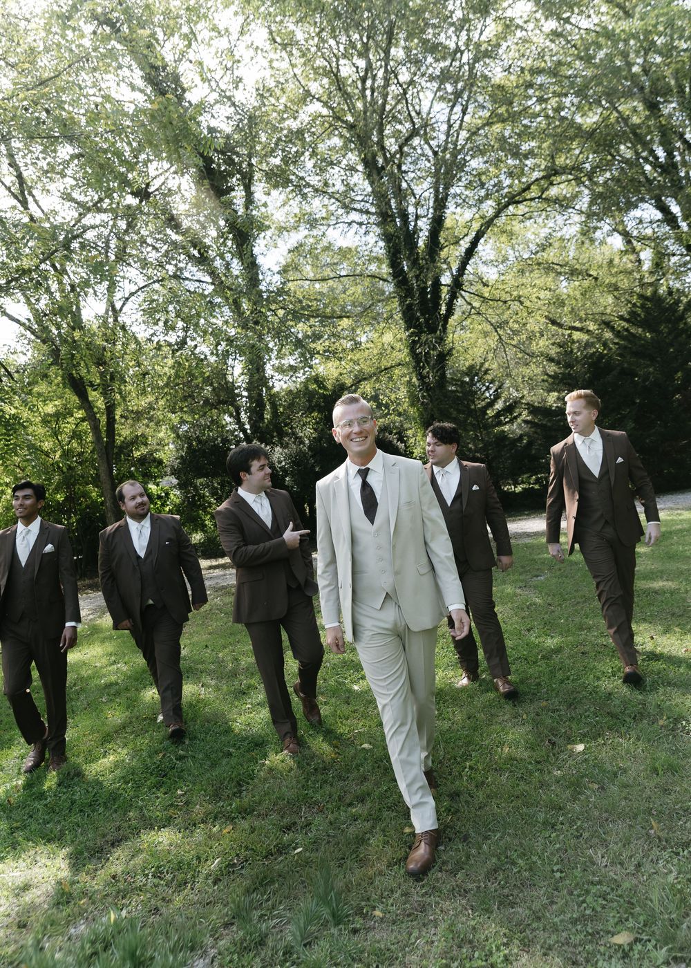 Groomsmen in dark suits walk behind the groom in a light gray suit on a grassy path surrounded by trees.