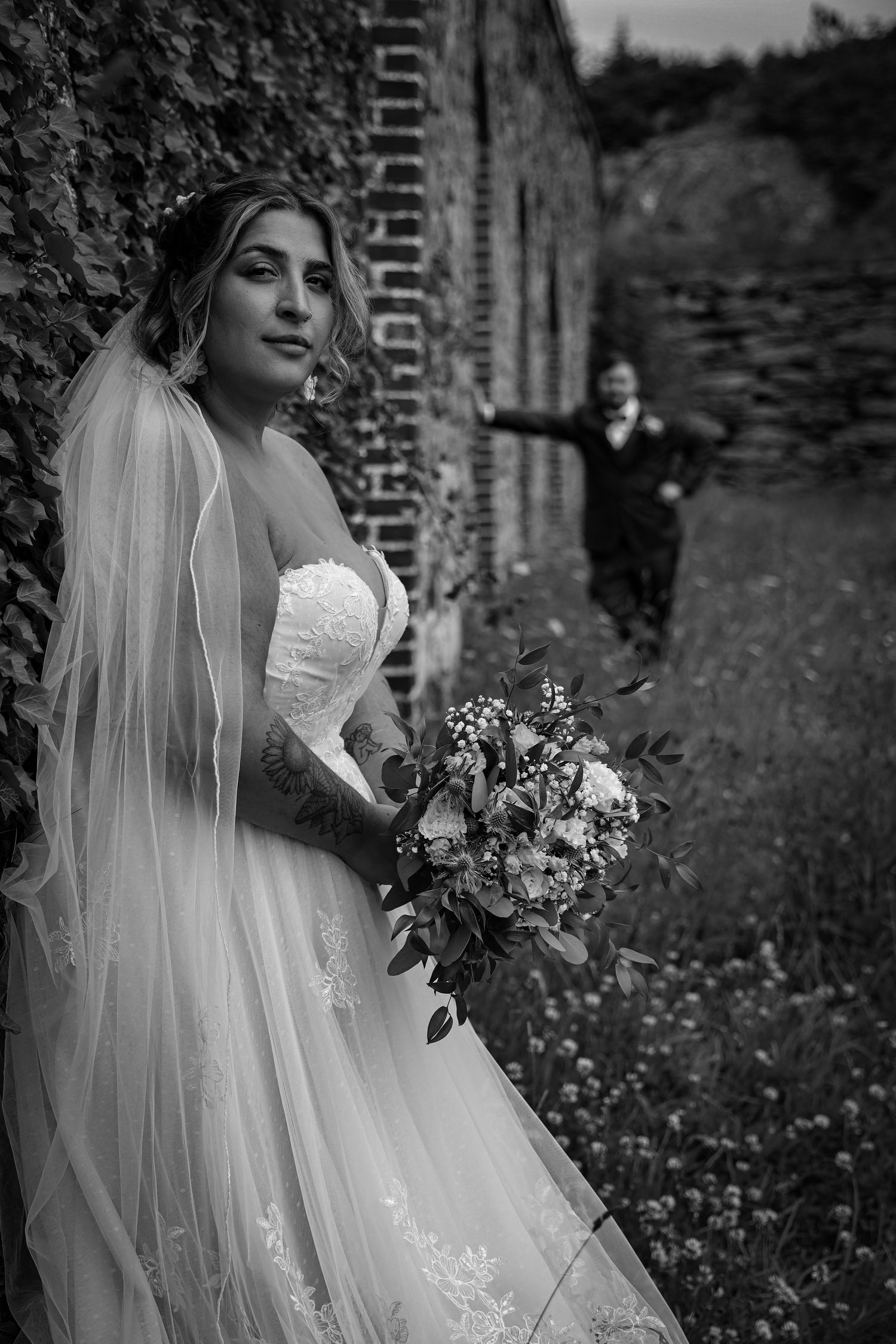 Bride in strapless lace gown with veil holds bouquet against brick wall as groom approaches.