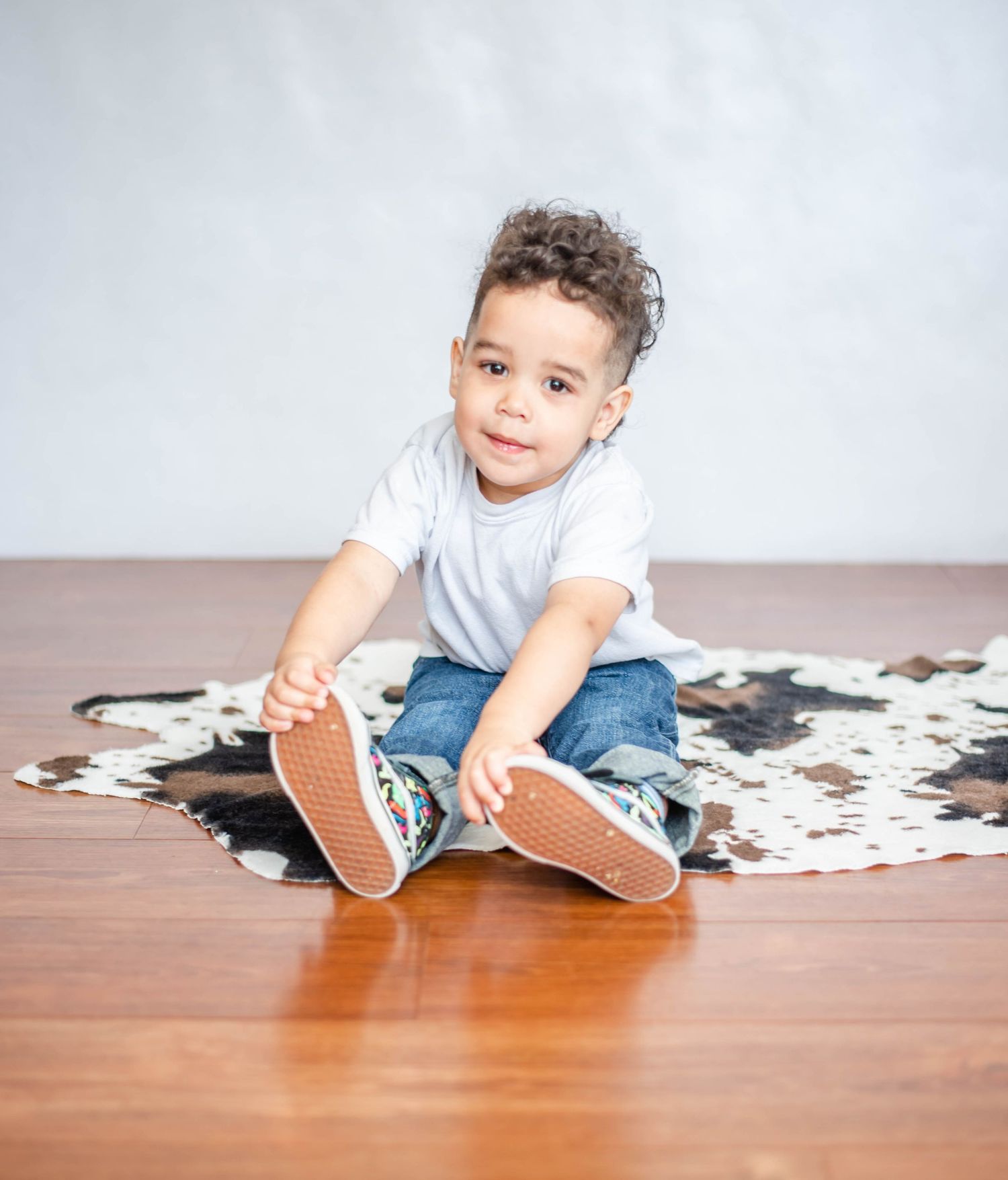 Child playing with shoes while sitting on cowhide print rug on wooden floor.