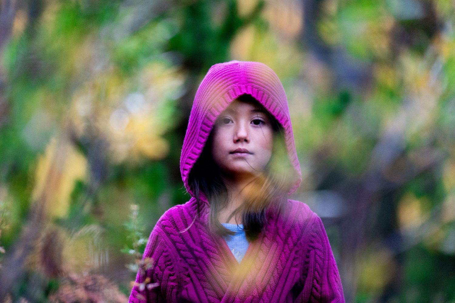 A person in a bright pink hoodie poses mysteriously in a blurred nature setting.
