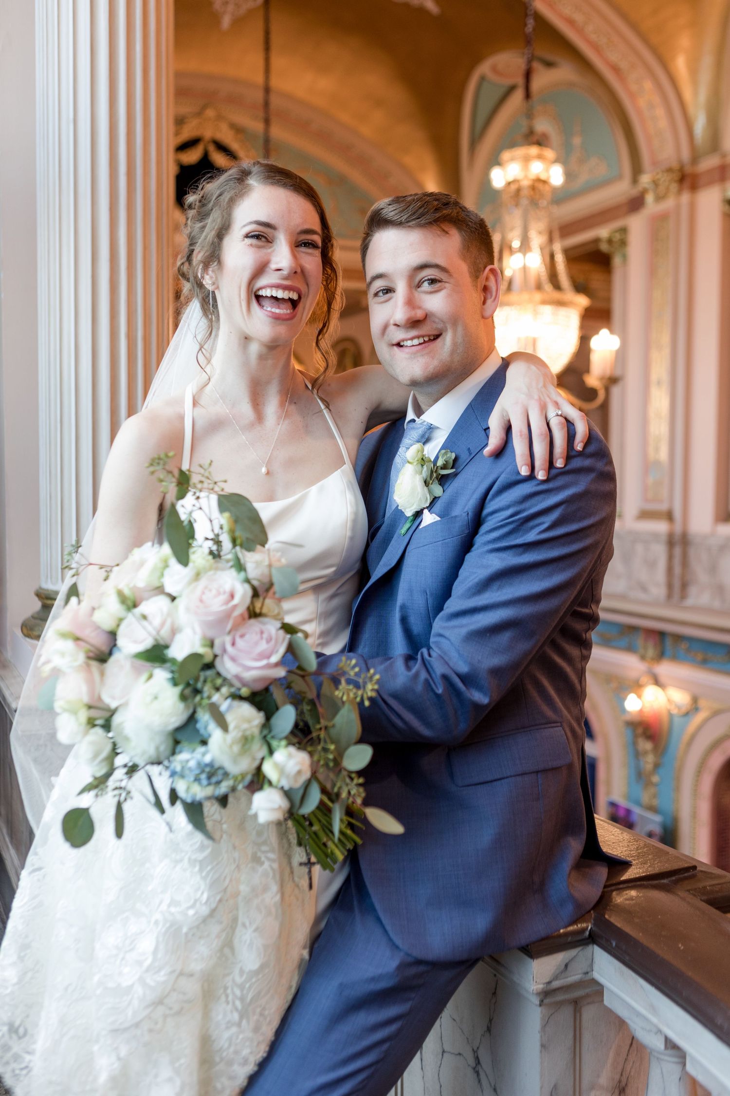 Joyful wedding couple shares candid laughs in an elegant ballroom with ornate columns and chandeliers.