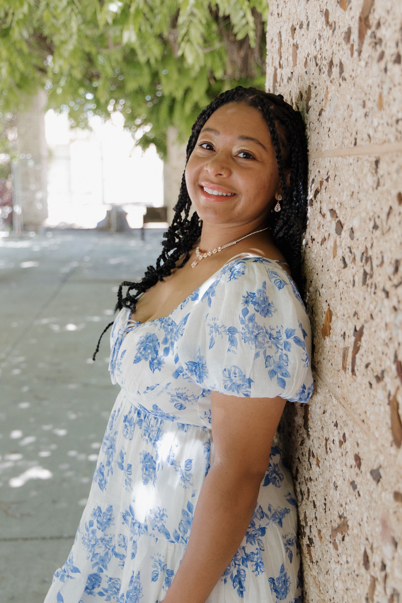 Woman in floral blue and white dress poses against stone wall with trees in background.